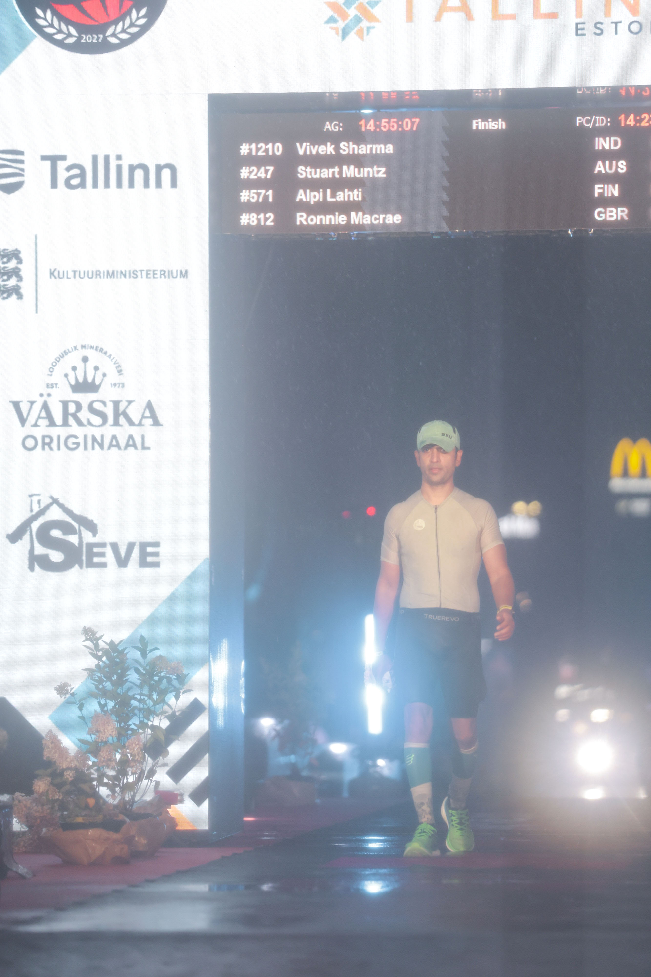 IRONMAN Tallinn finish scoreboard showing #1210 Vivek Sharma IND as Vivek walks through the finish arch at night